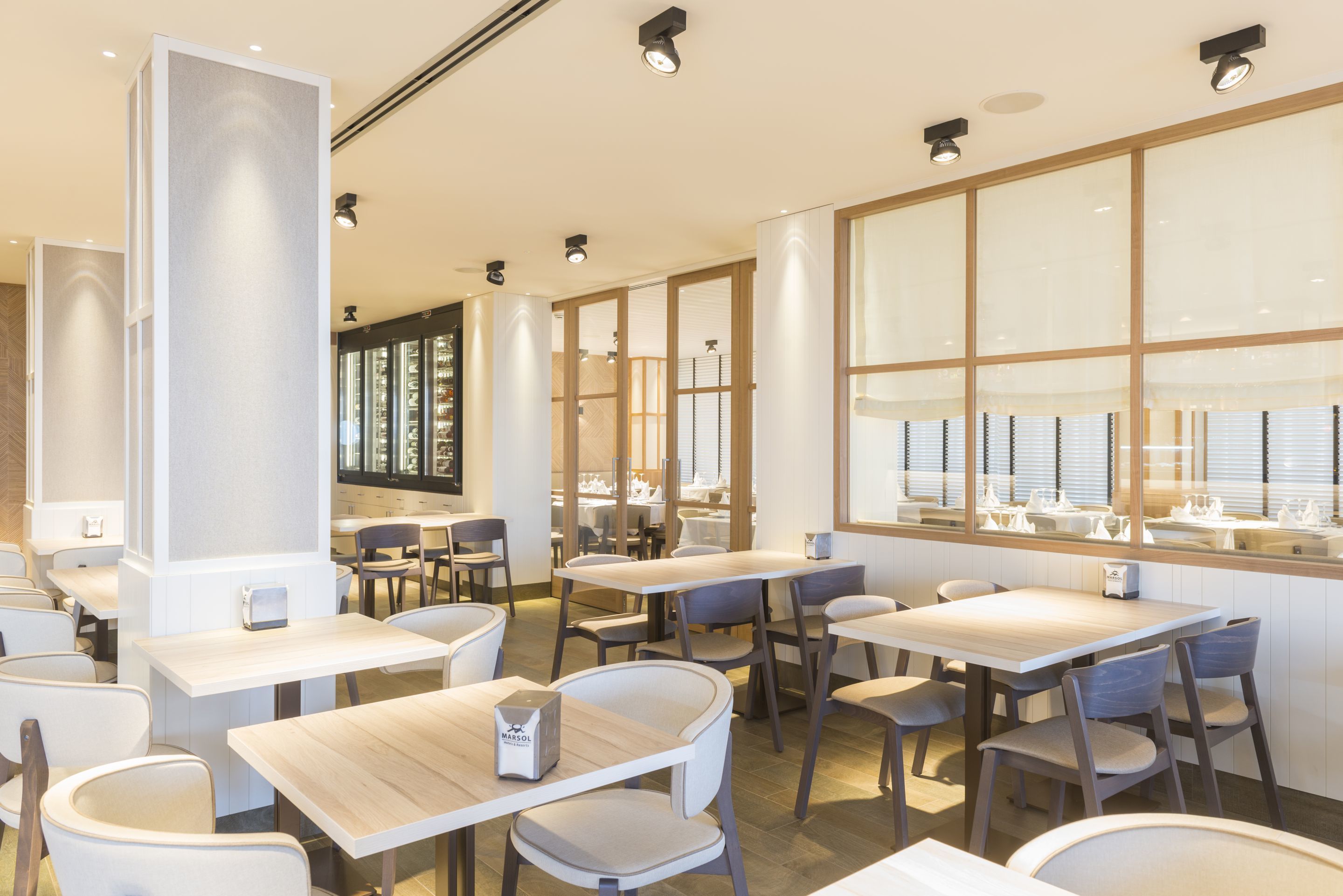 Ein modernes Restaurantambiente mit Holztischen und -stühlen, großen Fenstern und eingelassener Beleuchtung.