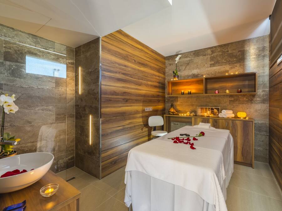 Un spa moderno con un amplio lavamanos blanco, paneles de madera, una camilla de masaje y un pequeño estante con velas.