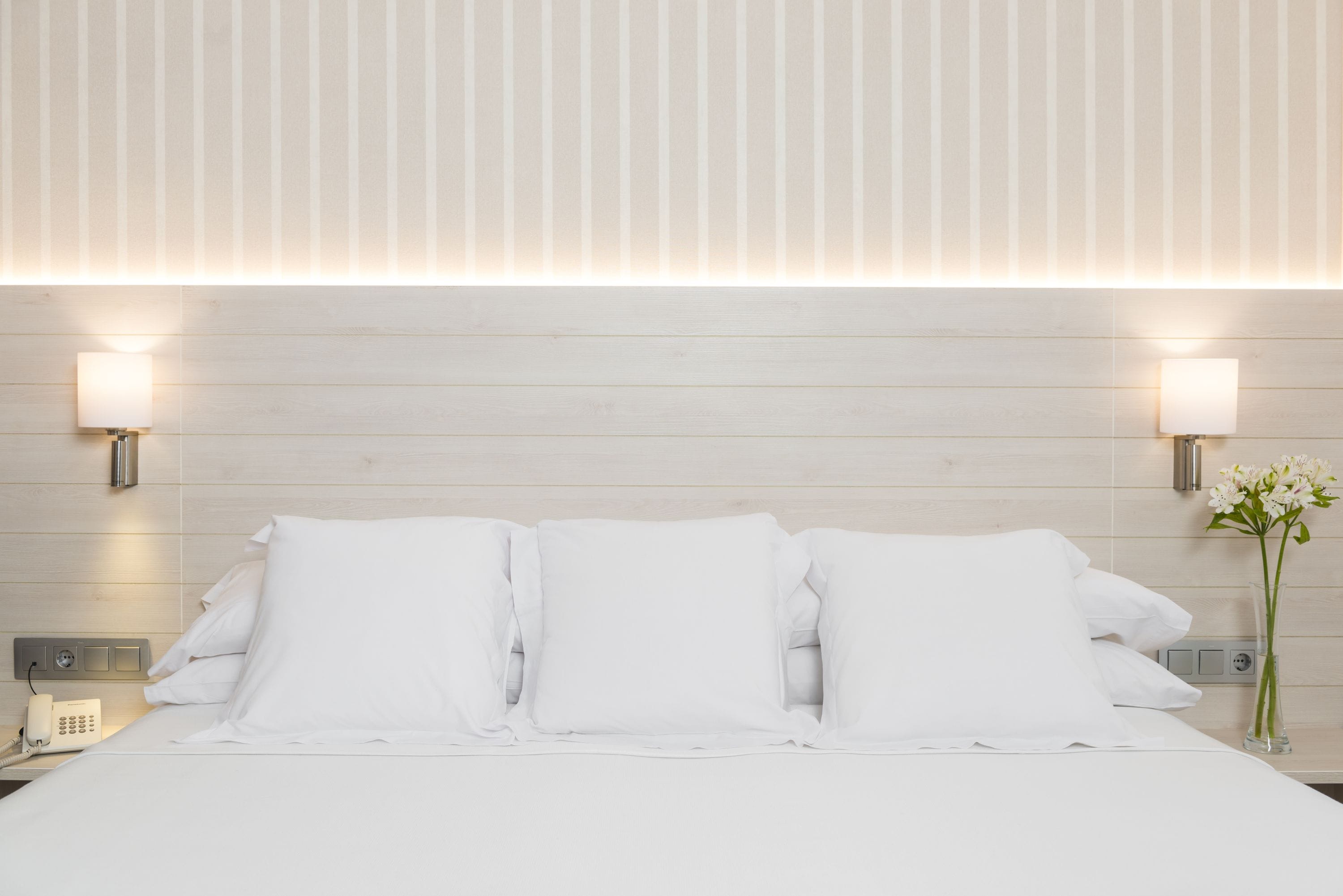 Een hotelkamer met een opgemaakt bed, twee bedlampjes, een telefoon op het nachtkastje en een vaas met bloemen op de bijzettafel.