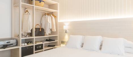 A hotel room with a neatly organized wardrobe, a bed with white linens, and a bedside table with a lamp.