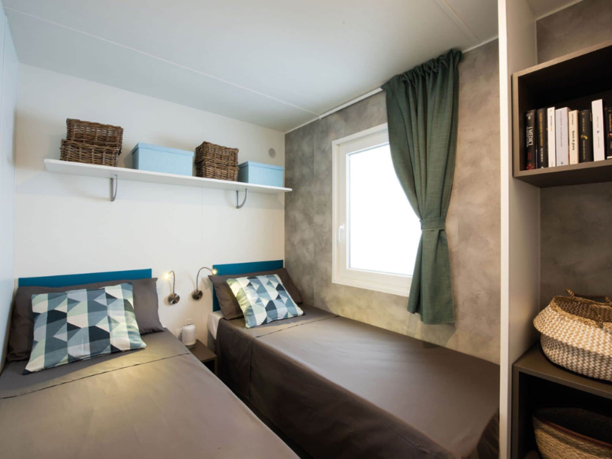 Une chambre avec deux lits, une fenêtre avec des rideaux, des étagères avec des paniers et des lampes fixées au mur.