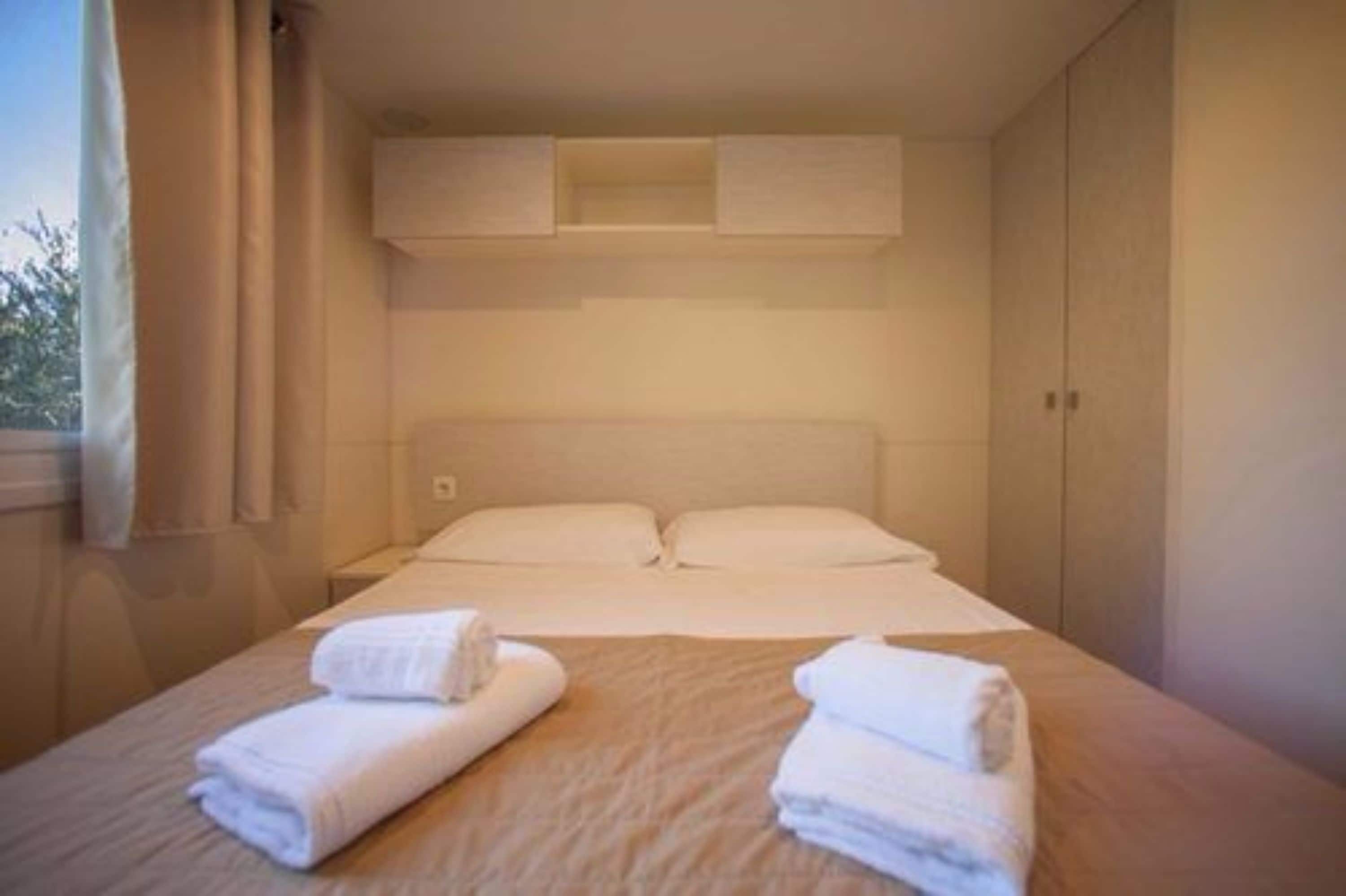 A hotel room with a bed, two pillows, a nightstand, and two rolled towels on the bed.