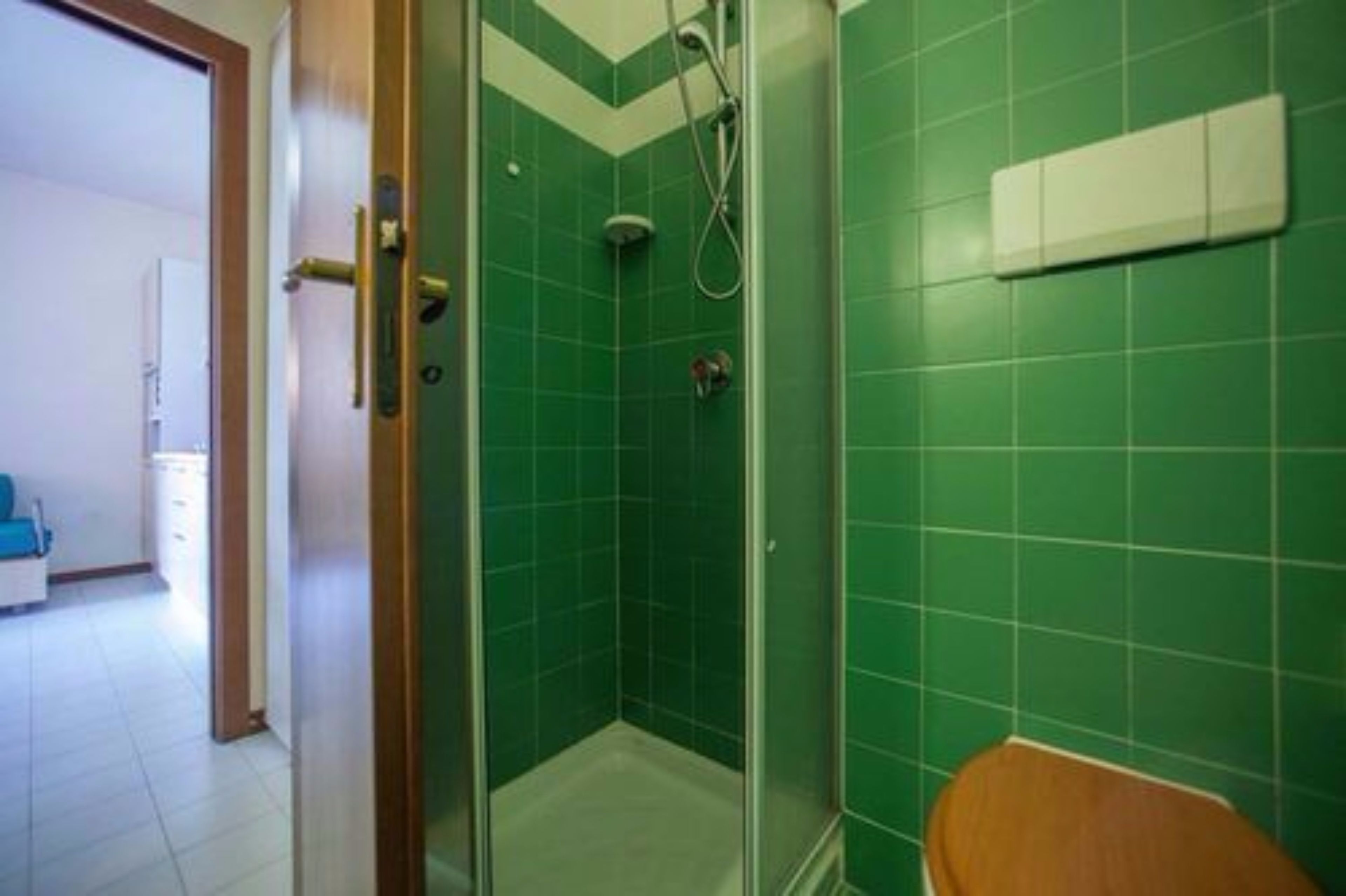 Un baño con baldosas verdes, cabina de ducha de vidrio y inodoro de madera.