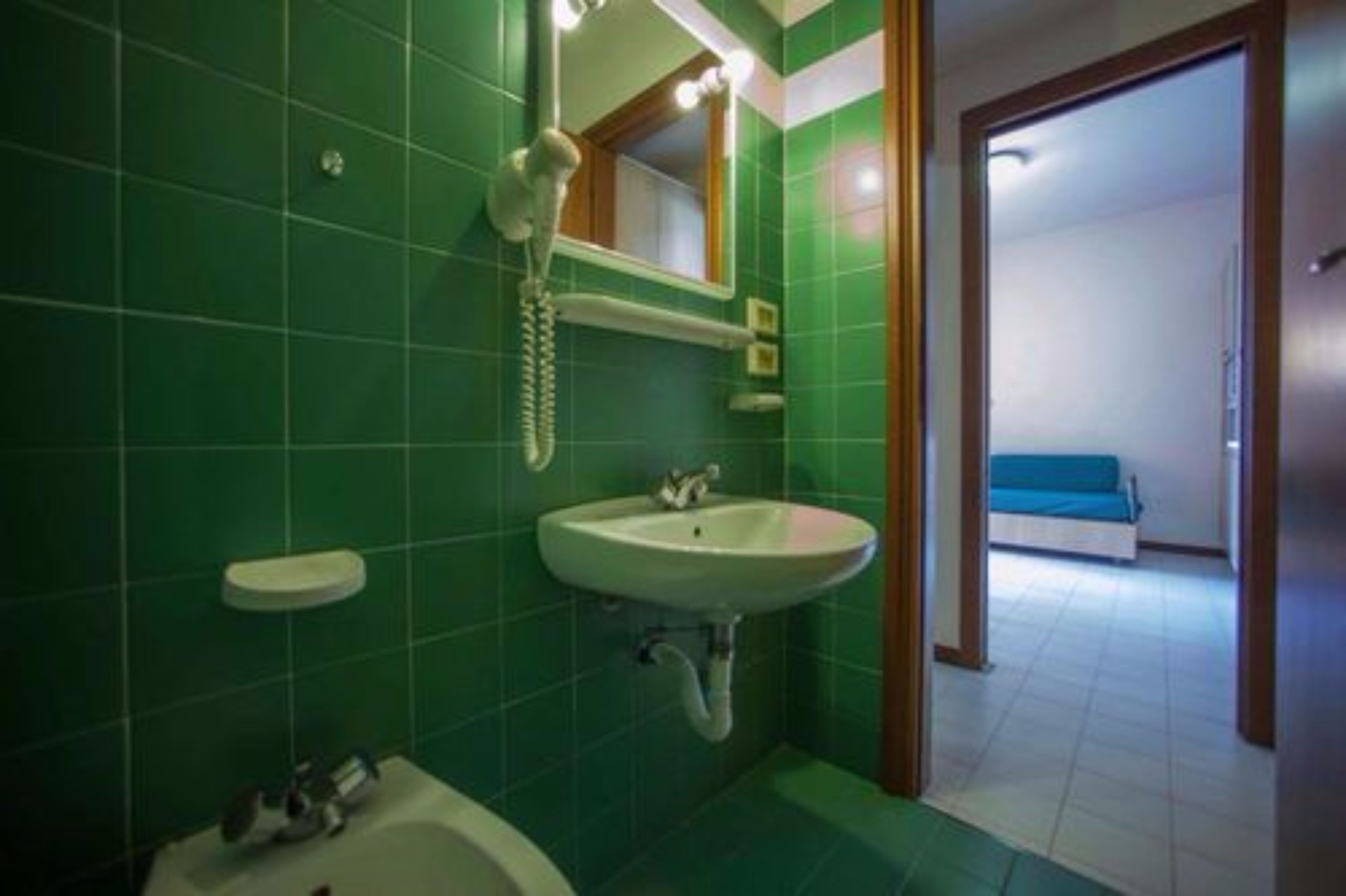 Une salle de bain avec des carreaux verts, un lavabo blanc, un miroir et un sèche-cheveux.