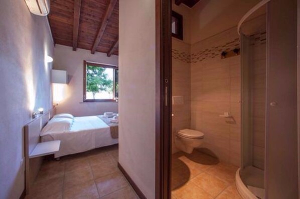 A bedroom with a bed, bedside table, and a window. Adjacent is a bathroom with a toilet, shower, and sink. - The Garda Village (Sirmione)