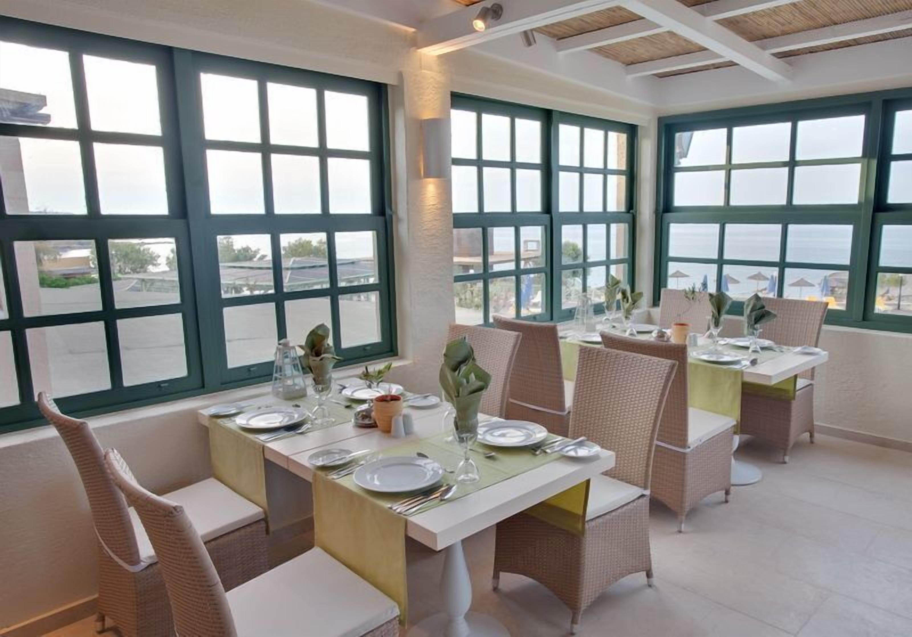 Un comedor con mesas dispuestas para cuatro, ventanas amplias con marcos verdes, sillas de mimbre y vistas al mar.