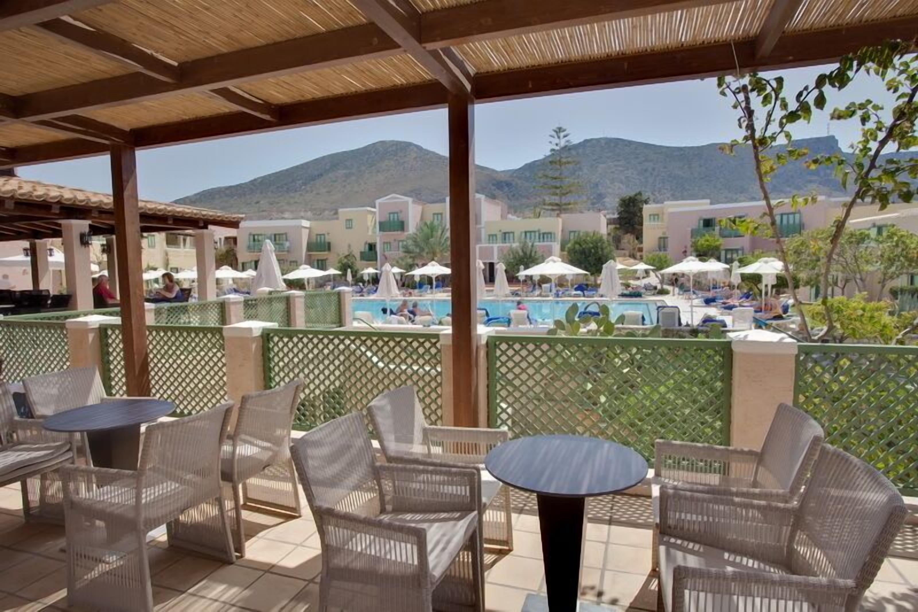 Ein überdachter Außenbereich mit Korbmöbeln, einem Pool und Gebäuden im Hintergrund.
