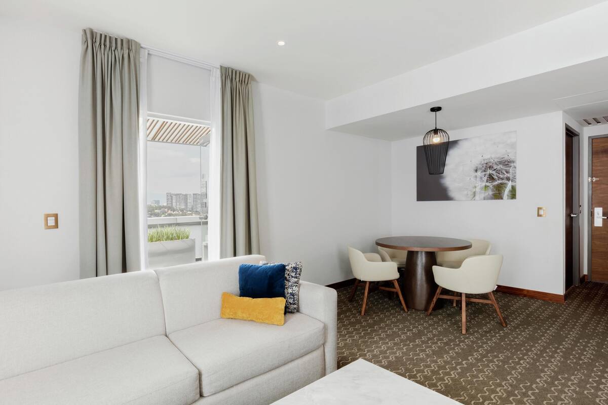 Habitación de hotel moderna con sofá, una mesa de comedor con sillas, una lámpara colgante y vistas a la ciudad.