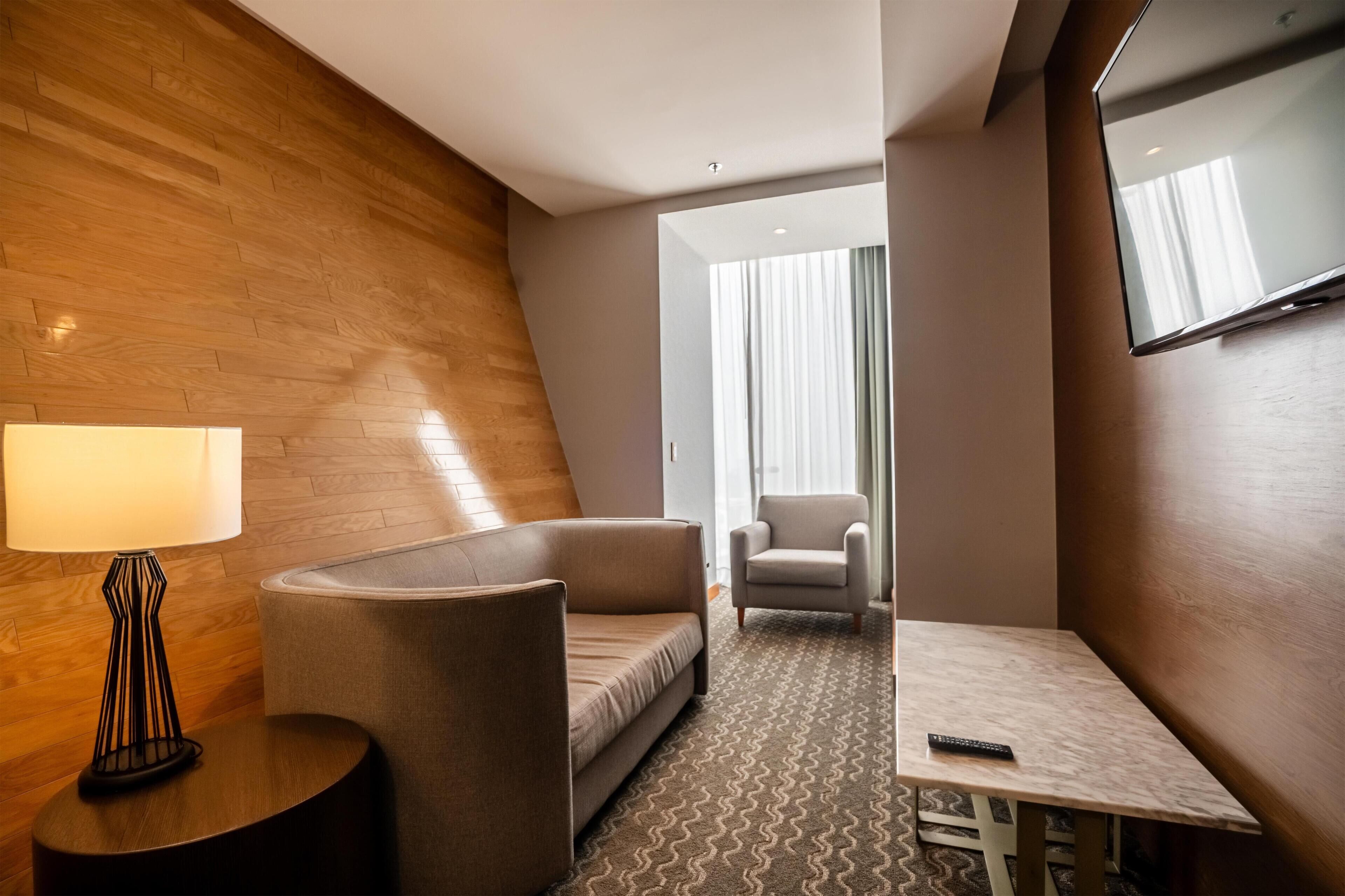 A hotel room with a sofa, armchair, small table, lamp, and a TV mounted on the wall.