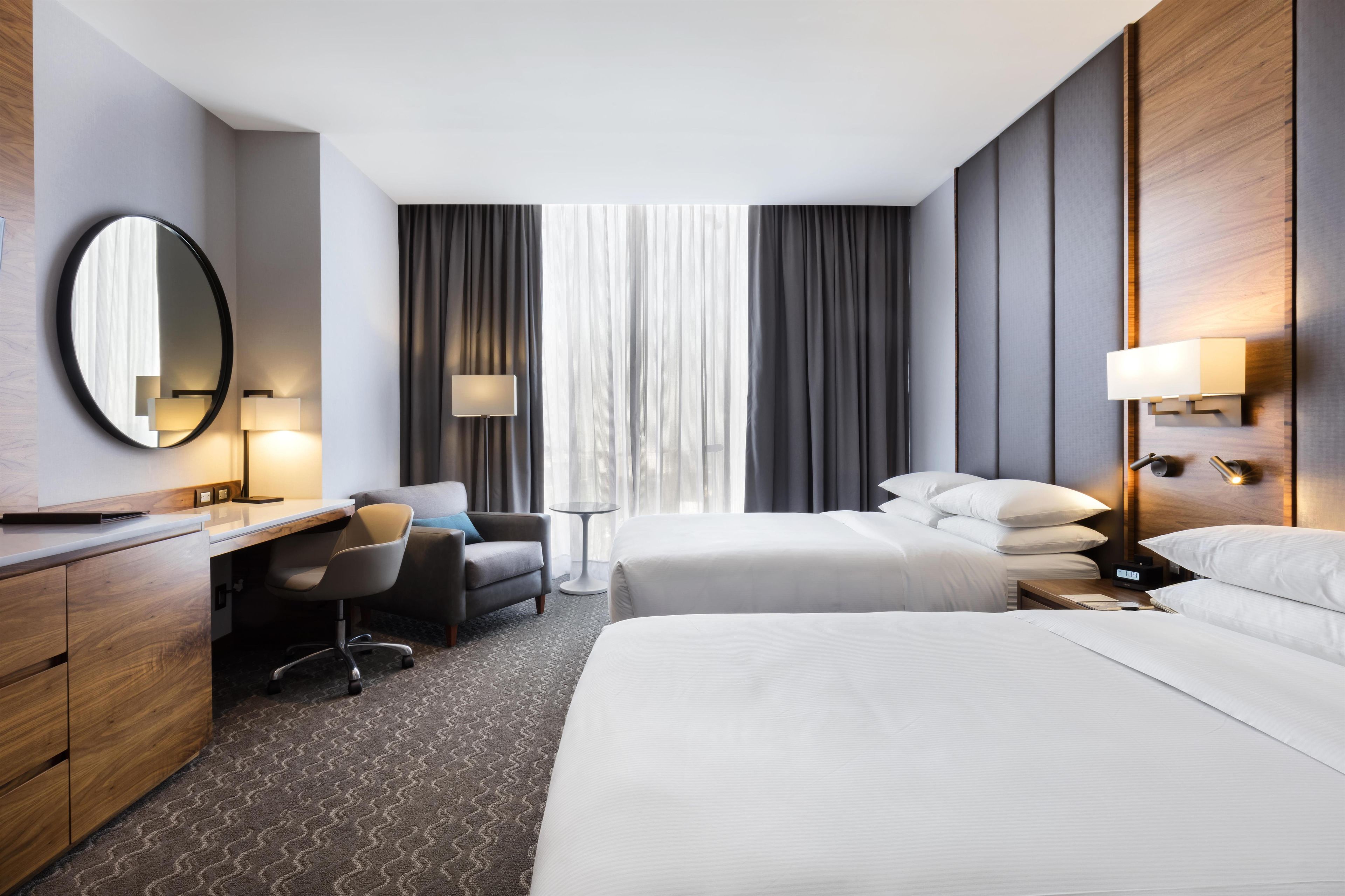 A hotel room with a desk, two beds, a chair, a mirror, and a window with curtains.