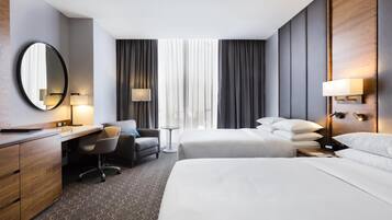A hotel room with a desk, two beds, a chair, a mirror, and a window with curtains.
