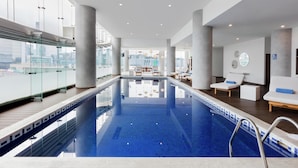 A modern indoor swimming pool with clear glass walls, lounge chairs, and a view of the cityscape. - DoubleTree by Hilton Hotel Mexico City Santa Fe (Mexico City)