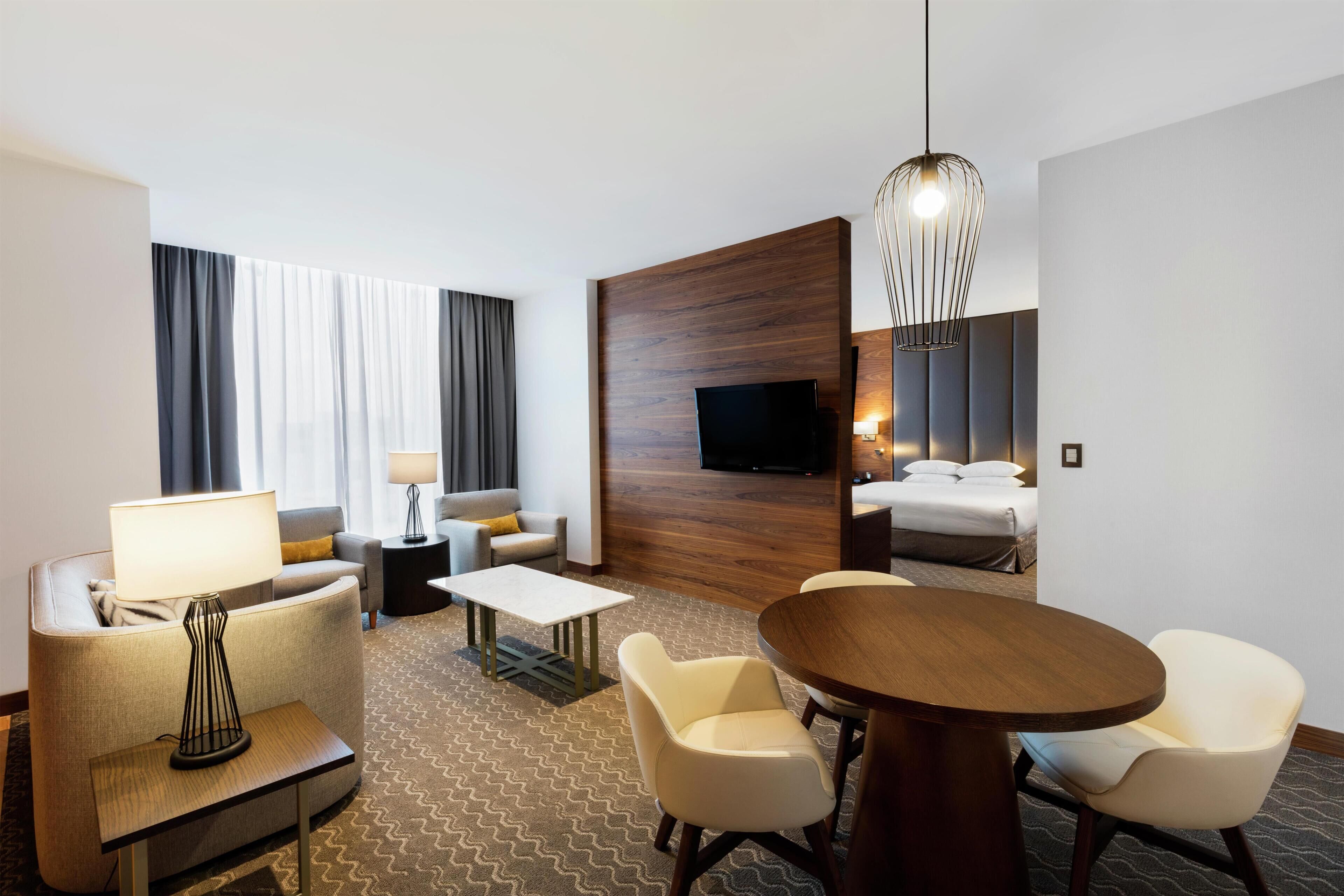 A hotel room with a sofa, armchair, dining table, TV, and a bed.