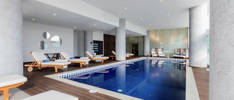 A modern indoor swimming pool with lounge chairs and a wooden deck.
