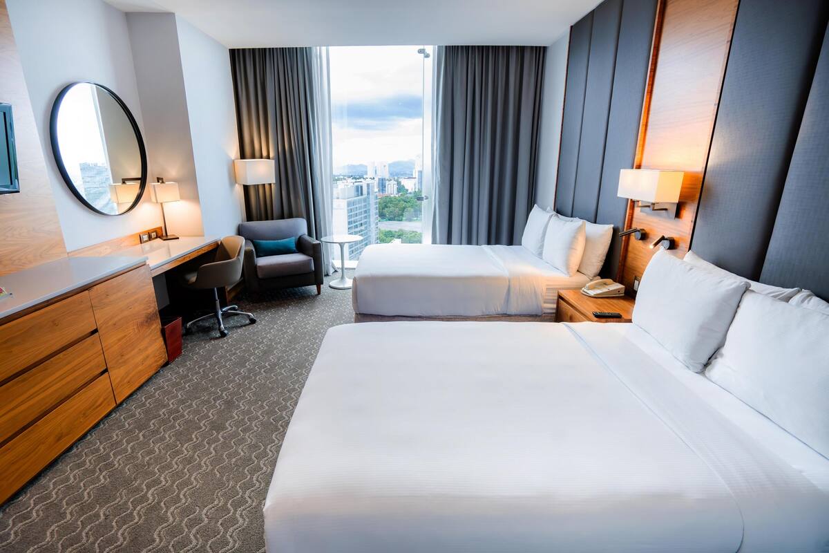 Habitación de hotel con dos camas, un escritorio, una silla, un espejo y vistas a la ciudad.