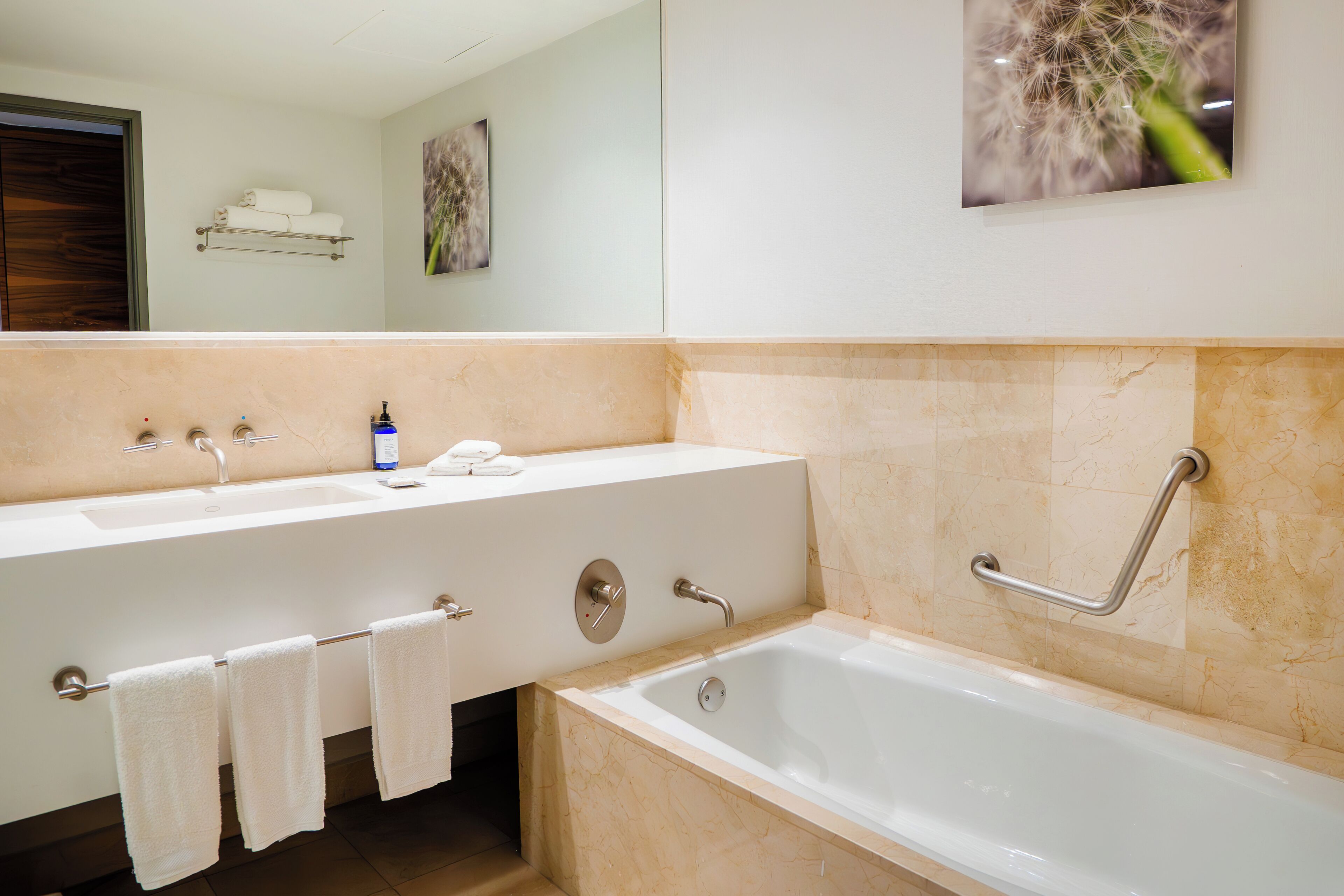 Une salle de bain avec un double lavabo, une baignoire et une barre d’appui. Des serviettes sont accrochées au mur et une étagère contient des articles de toilette.