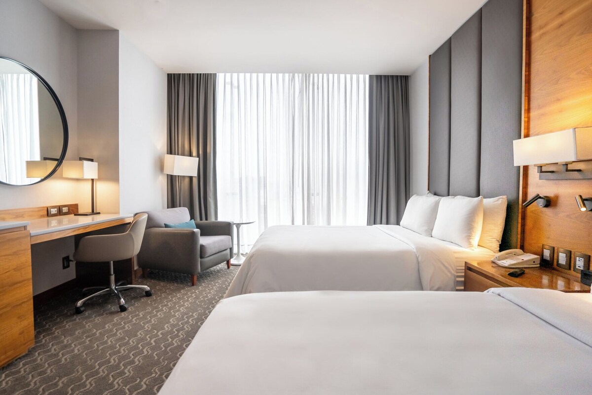 Une chambre d’hôtel avec deux lits, un bureau, une chaise, une table de chevet avec une lampe et une grande fenêtre avec des rideaux.