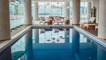 An indoor swimming pool with lounge chairs and sun loungers around it, large windows, and a view of the cityscape.
