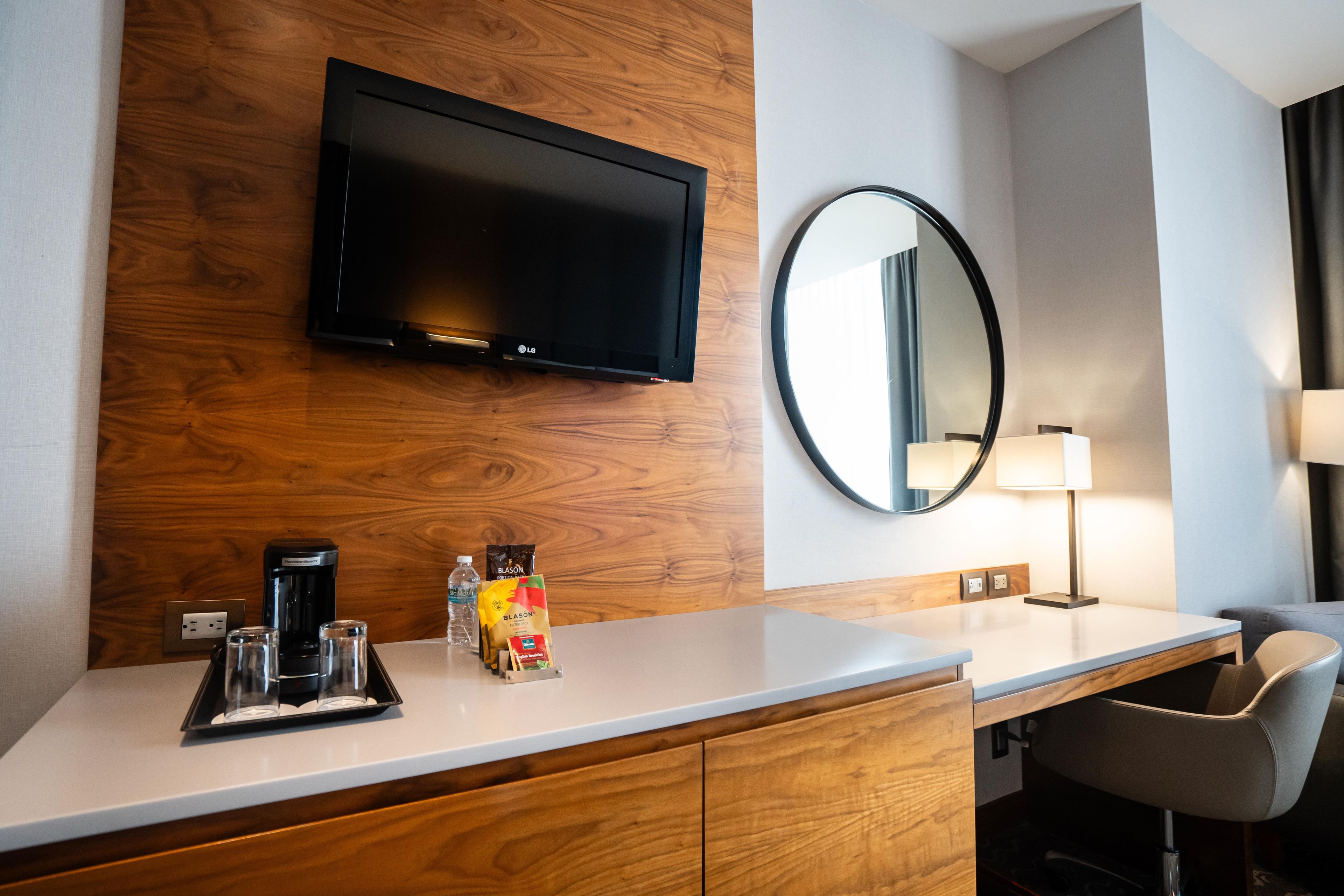 A hotel room with a flat-screen TV mounted on a wooden wall, a round mirror, a white countertop, a coffee maker, a water bottle, and a chair.