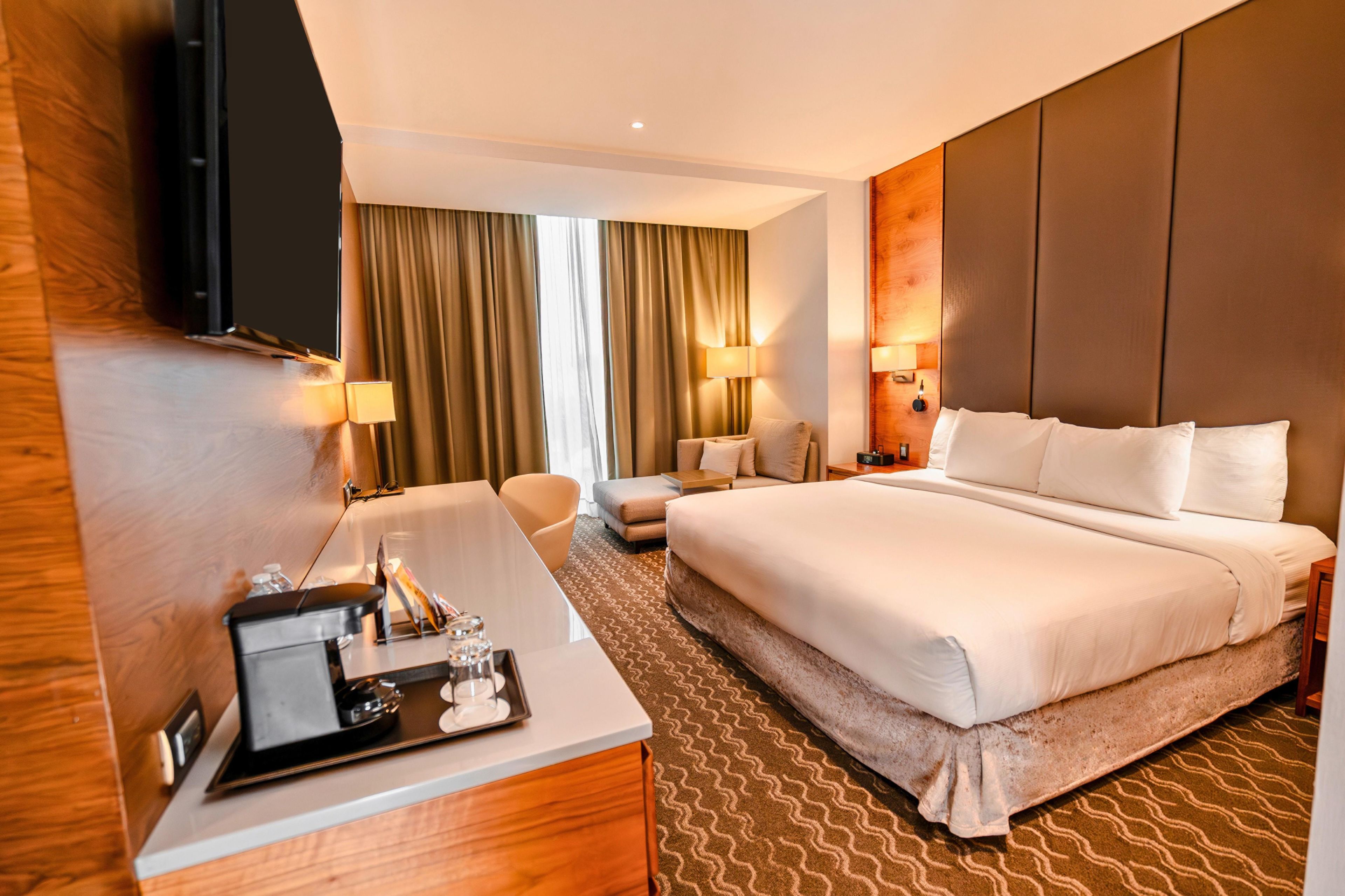 A hotel room with a large bed, a desk with a tray of amenities, a chair, a sofa, and a TV mounted on the wall.