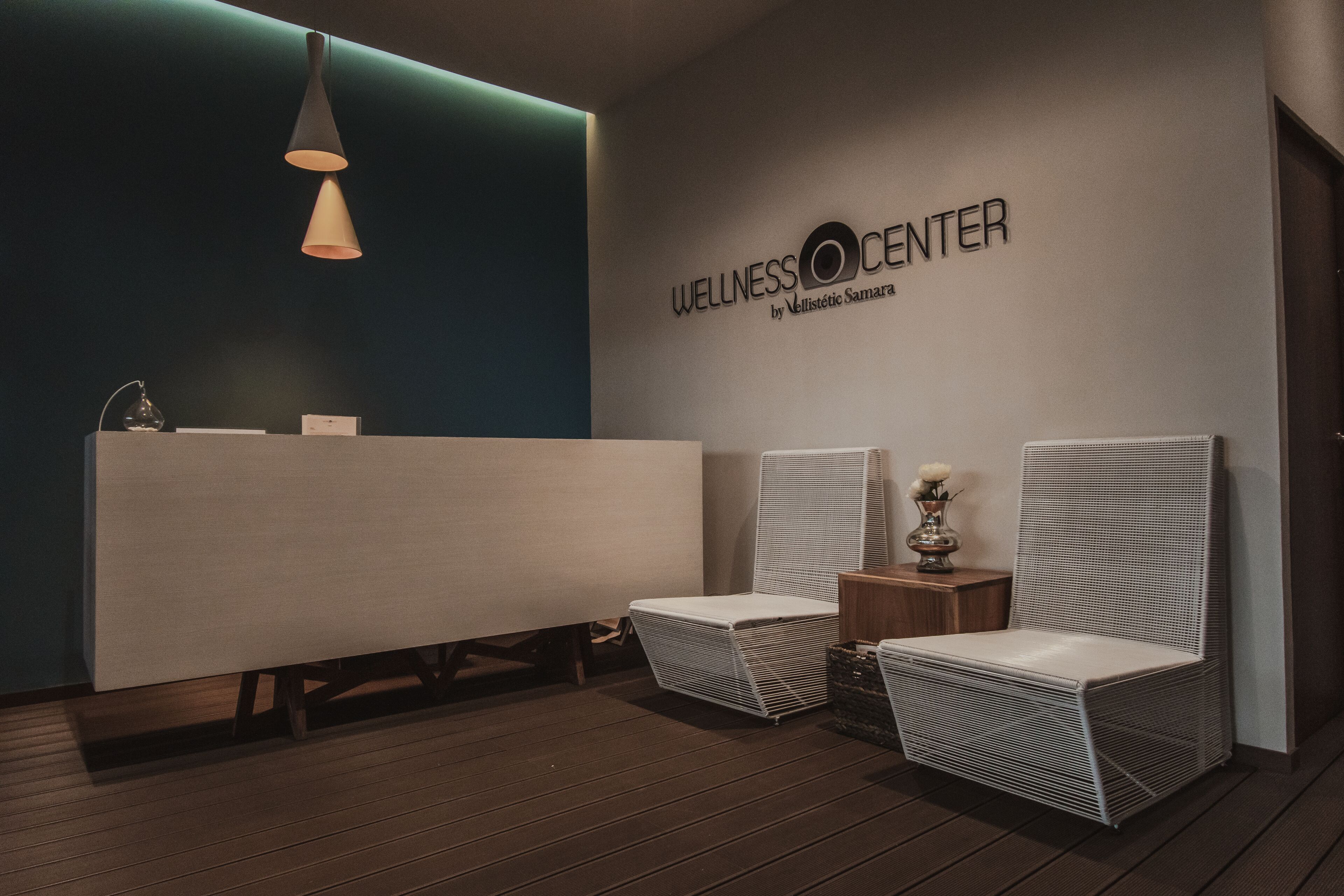 A wellness center with a reception desk, two wicker chairs, a small table with a vase, and a pendant light.