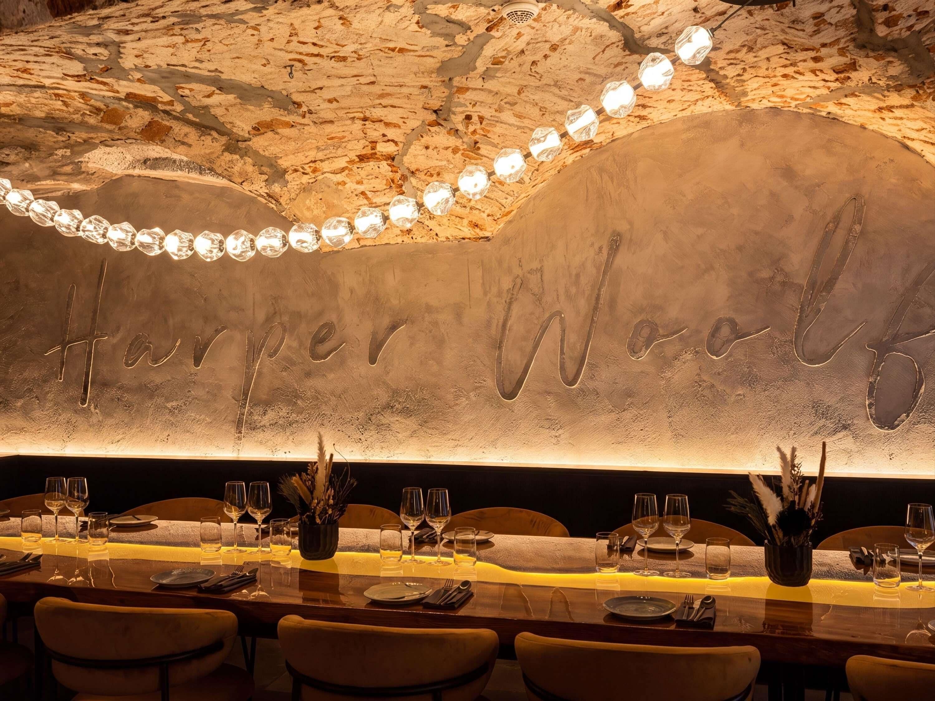 A restaurant interior with a long table set for dining, a wall with the name 'Harper Woolf' etched into it, and a ceiling with exposed rock-like texture.