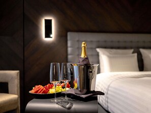 A neatly arranged room with a bed, a bedside table with a bottle of champagne, two glasses, and a fruit platter.