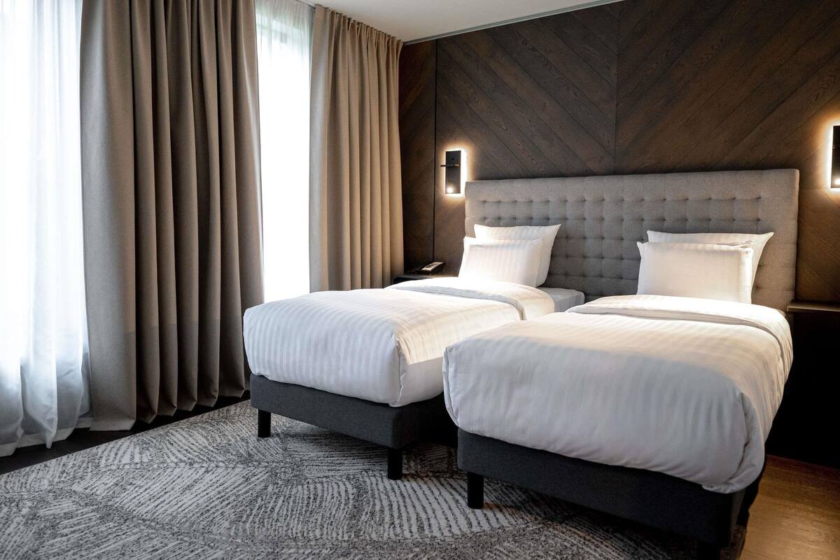 Ein Hotelzimmer mit zwei Betten, einem großen Fenster mit Vorhängen, einem modernen Kopfteil und einem gemusterten Teppich.