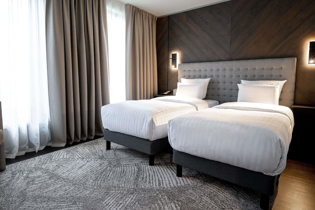 Ein Hotelzimmer mit zwei Betten, einer Wand in dunklem Holzvertäfelung und einem großen Fenster mit Vorhängen.