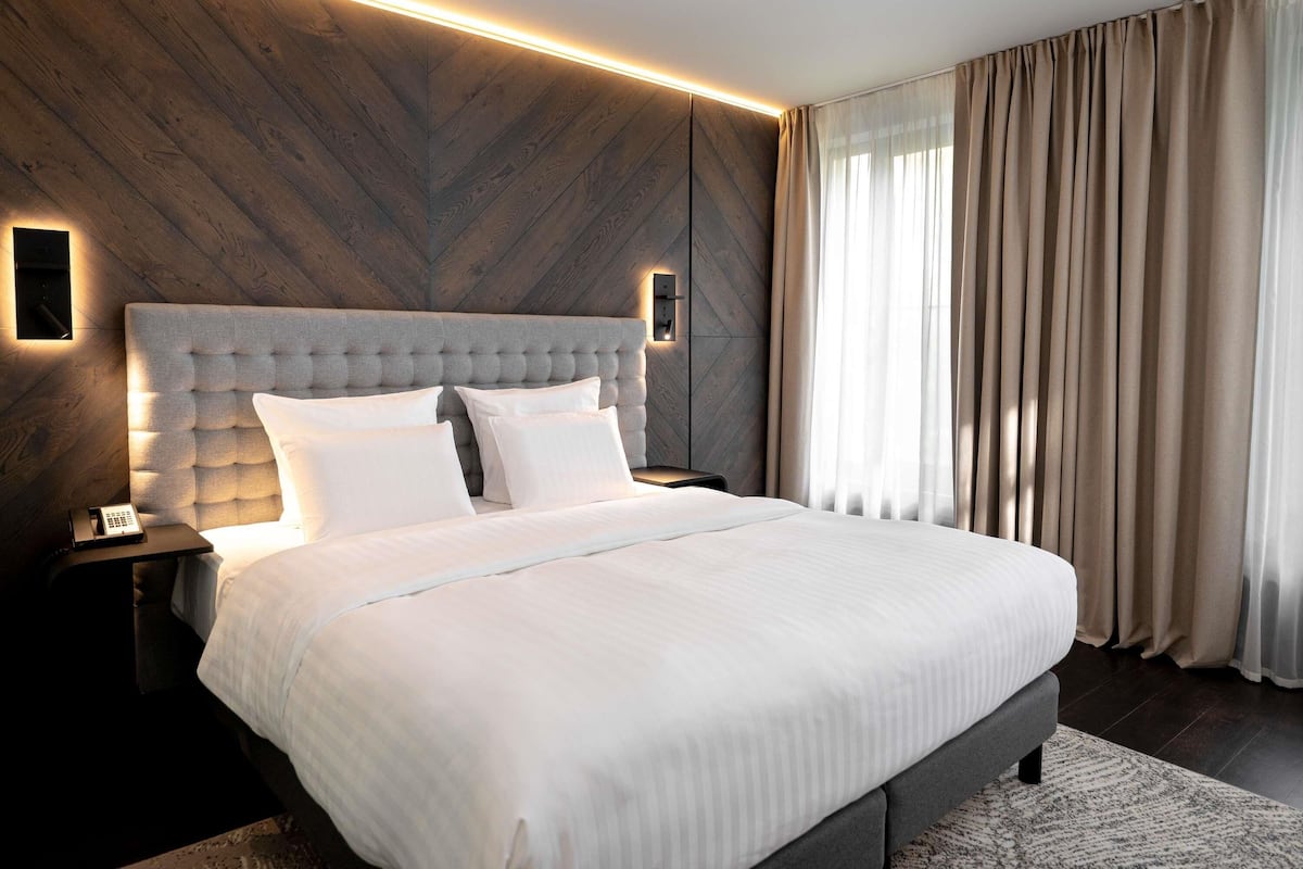 Ein modernes Hotelzimmer mit einem großen Bett, einem Nachttisch, einer Wandleuchte und einem Fenster mit Vorhängen.
