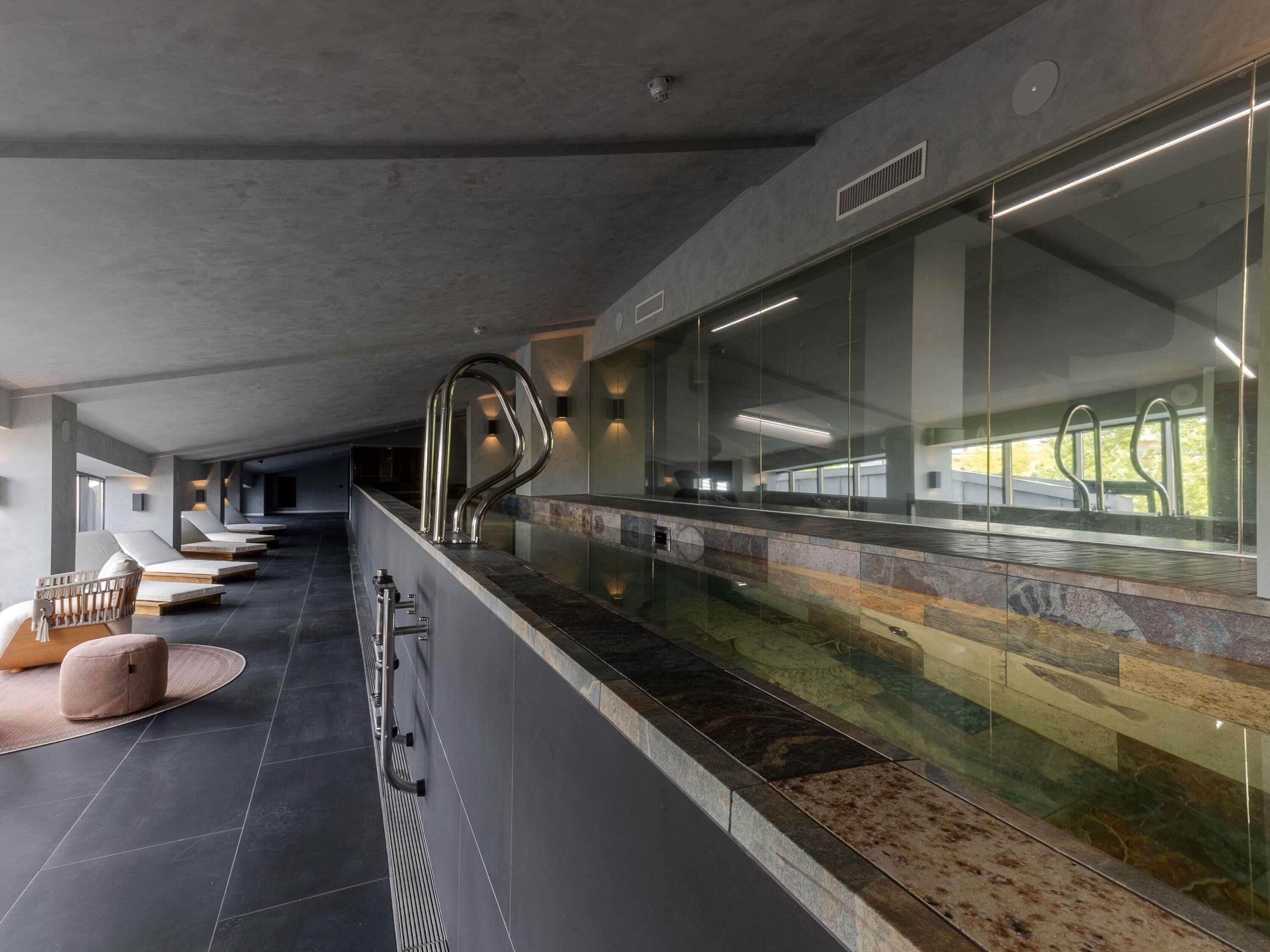 Un espace piscine intérieur moderne, doté d’un long banc, d’un mur en verre et d’une rambarde en métal.