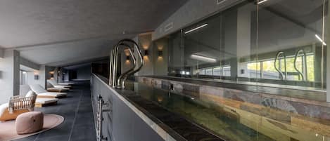 Un área de piscina interior moderna con un banco alargado, una pared de vidrio y una barandilla metálica.