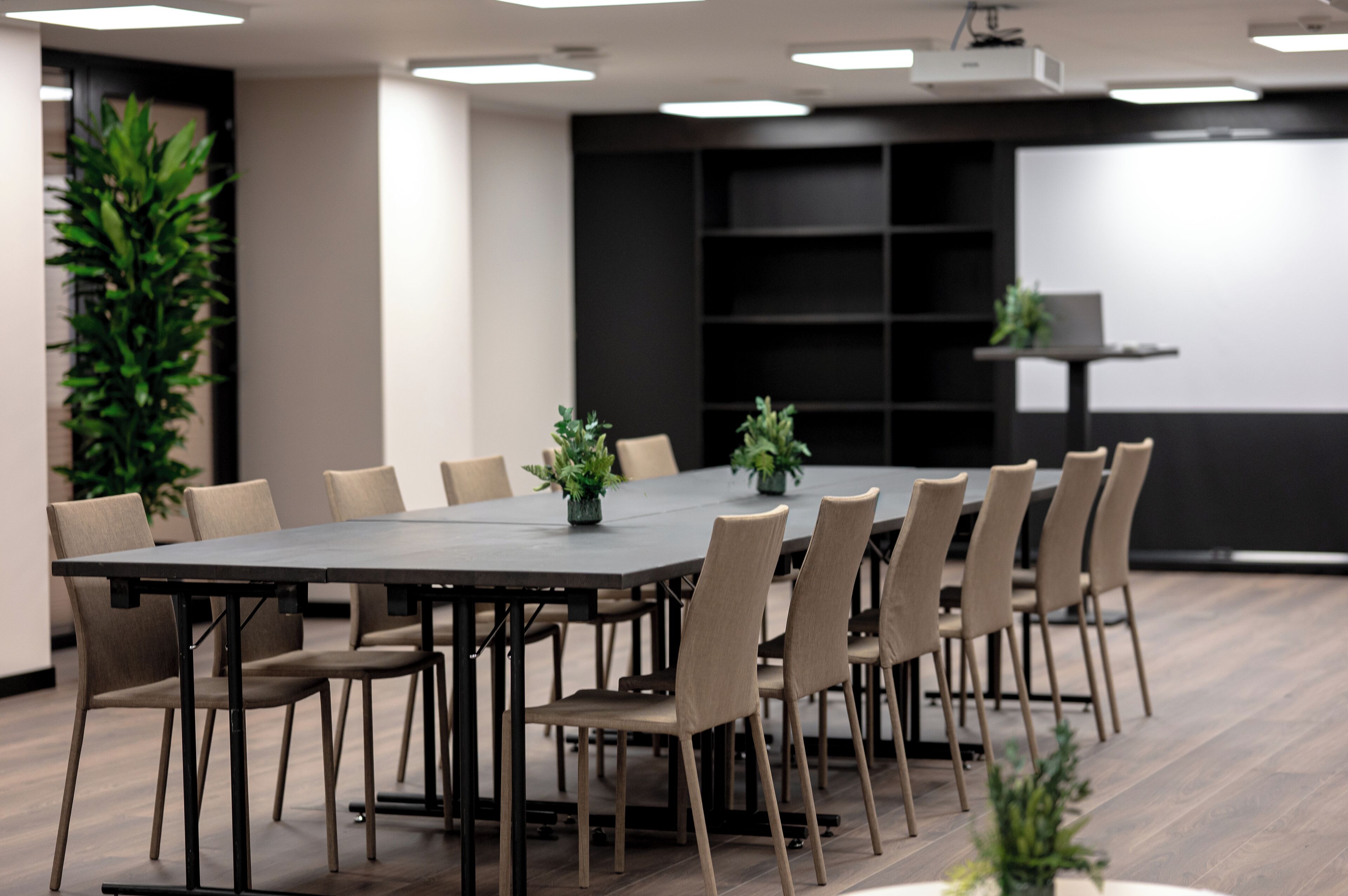 A conference room with a rectangular table surrounded by chairs, a projector screen, and potted plants.
