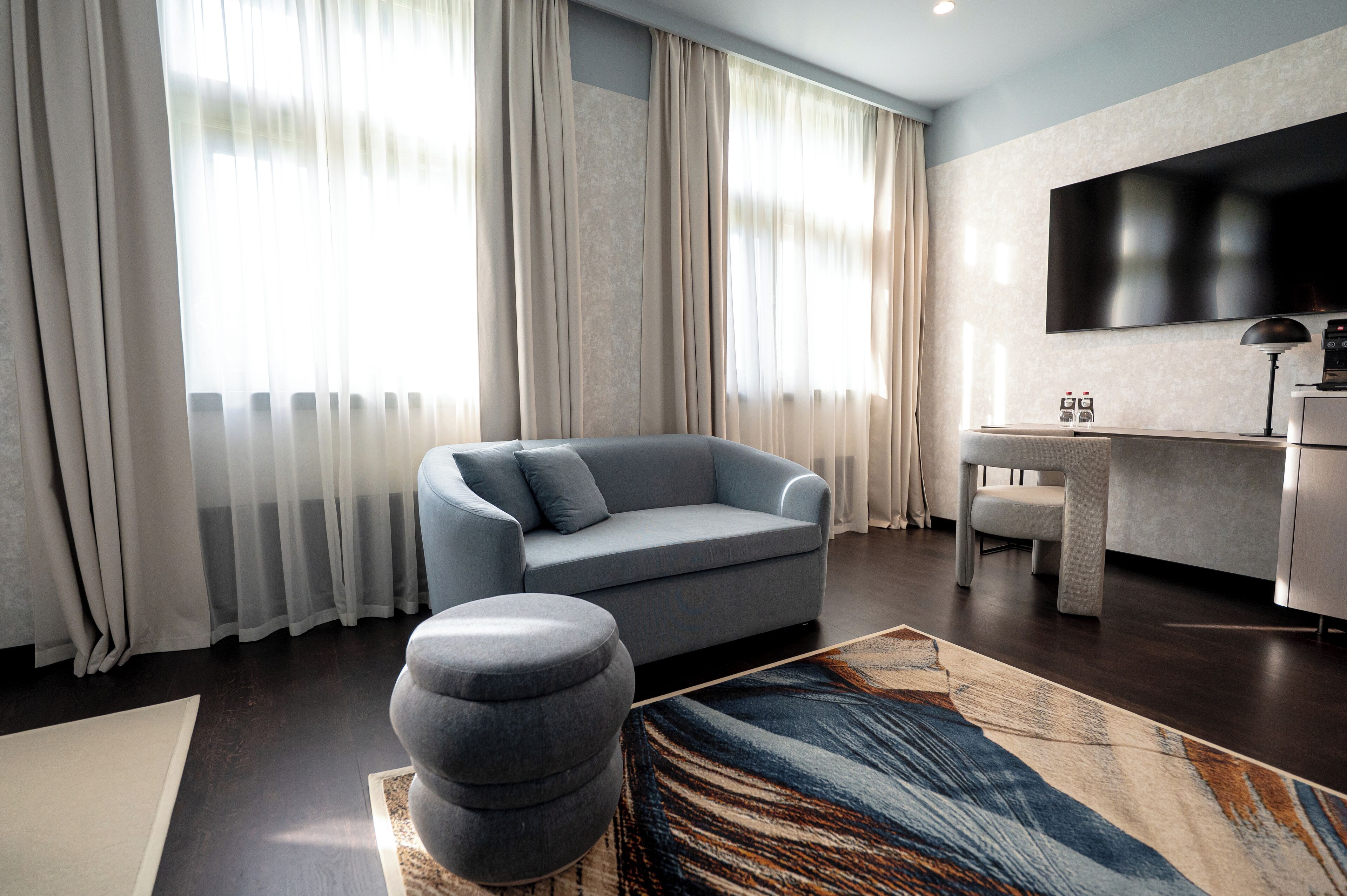 A modern living room with a grey sofa, a matching ottoman, a flat-screen TV mounted on the wall, and a small table with two glasses.