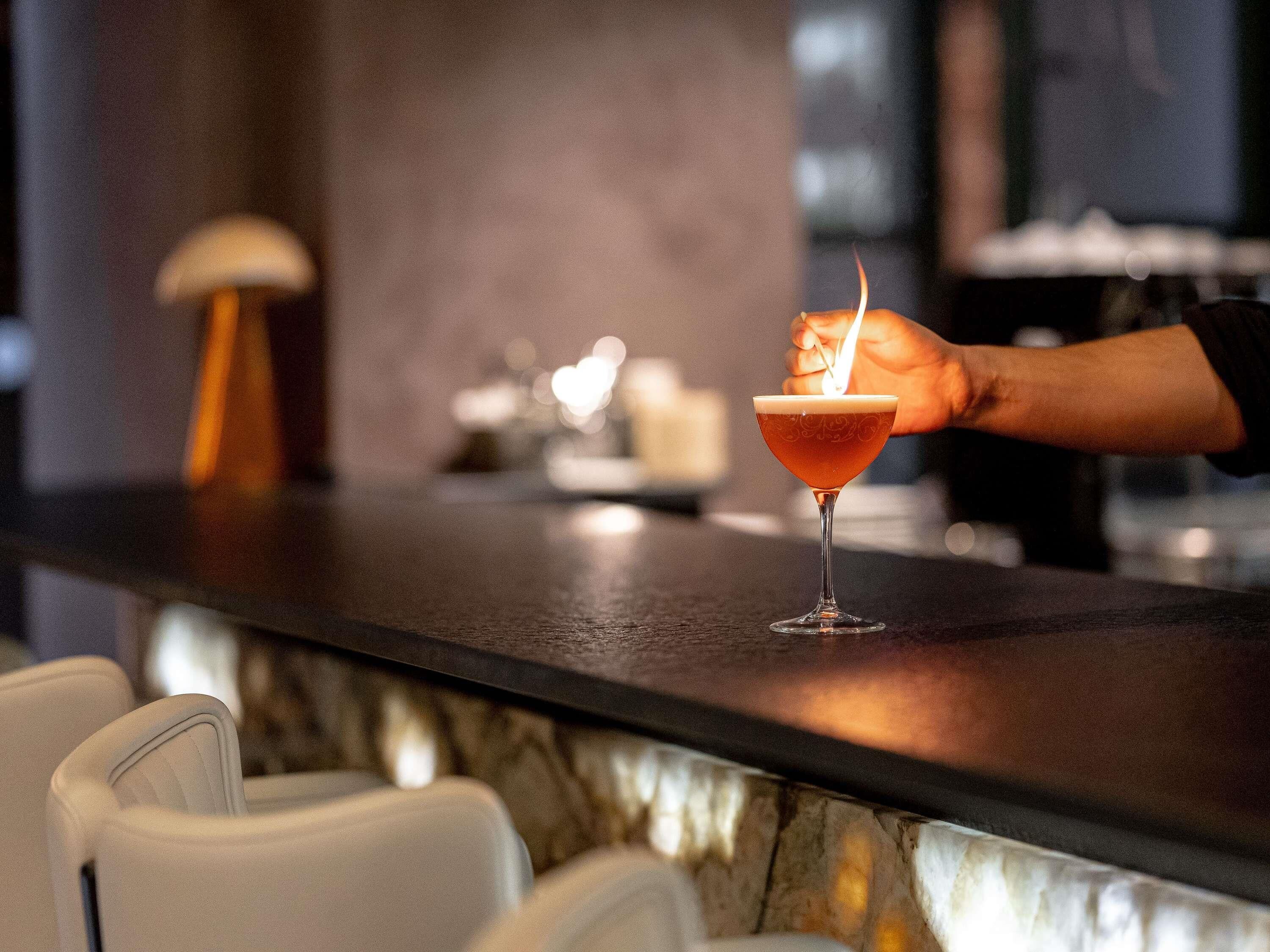 Un bar con un cóctel en una copa de martini, una mano sosteniendo un encendedor, y un fondo desenfocado con una lámpara y sillas.