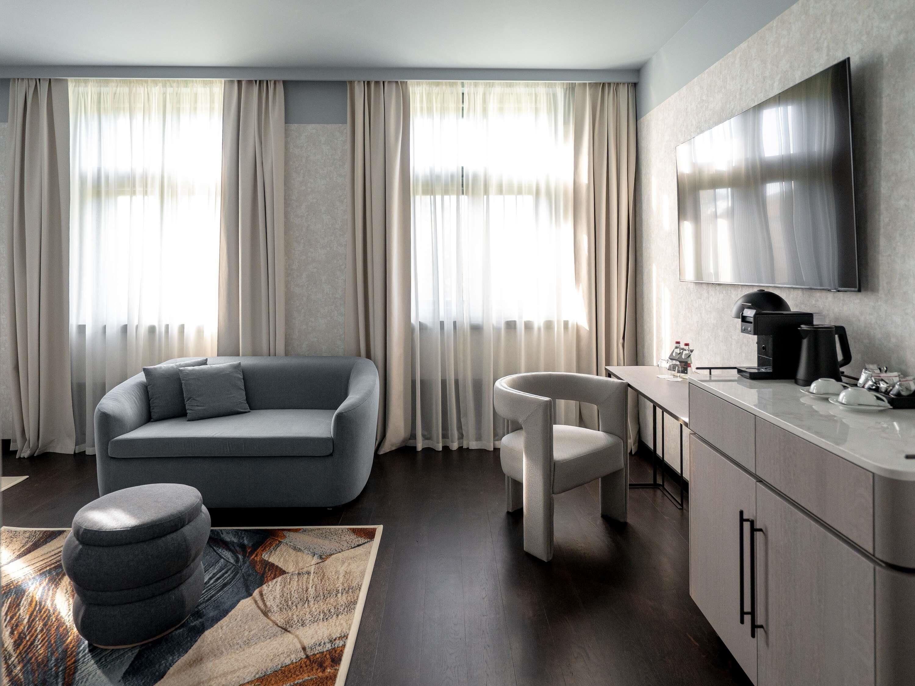 Una sala moderna con un sofá gris, un otomano redondo pequeño, una silla blanca, un área de cocina con una cafetera y un televisor de pantalla plana montado en la pared.