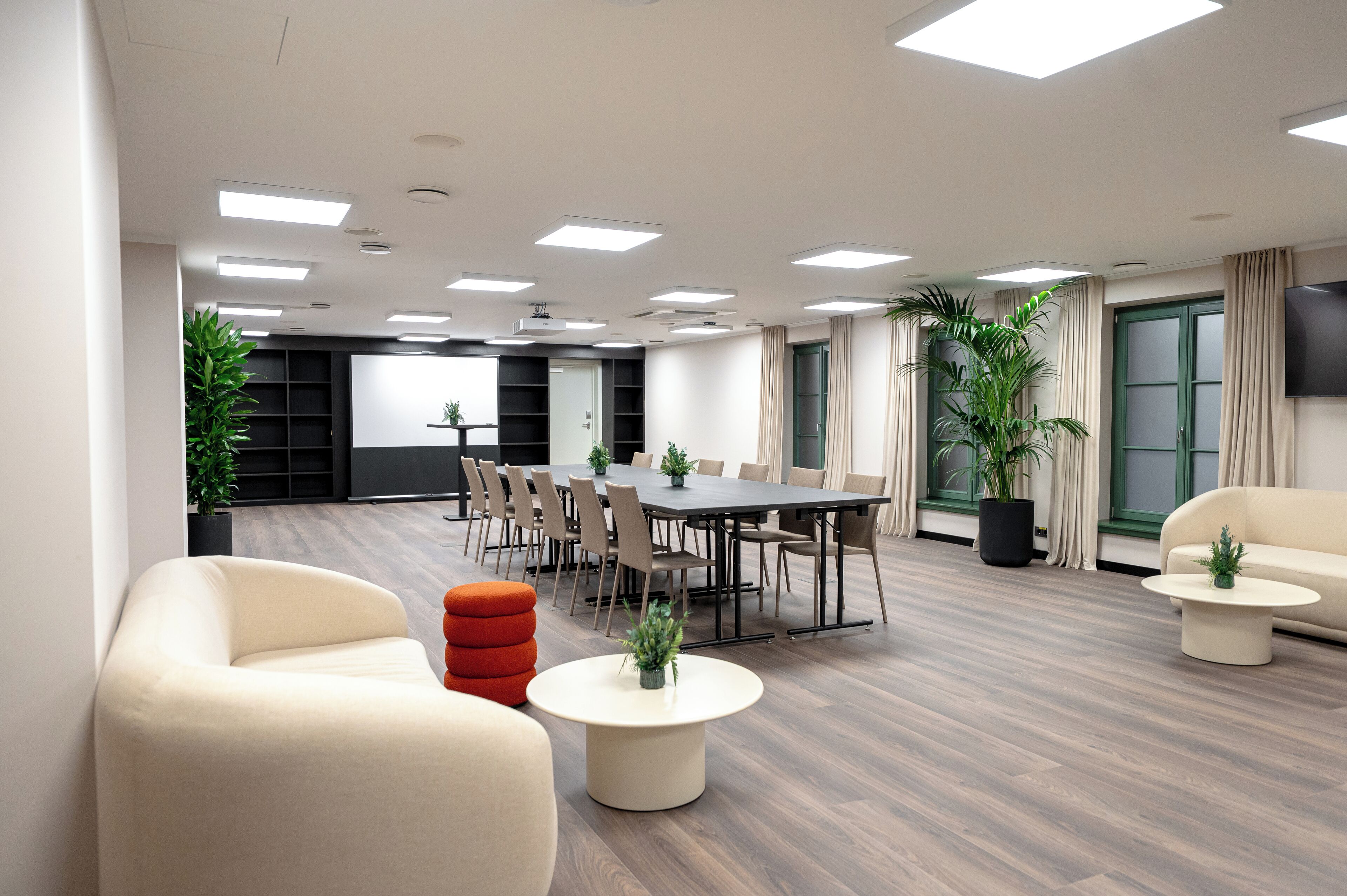 A modern conference room with a large table, chairs, and a sofa. There are potted plants and a television on the wall.