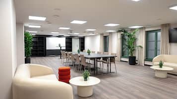 A modern conference room with a large table, chairs, and a sofa. There are potted plants and a television on the wall.