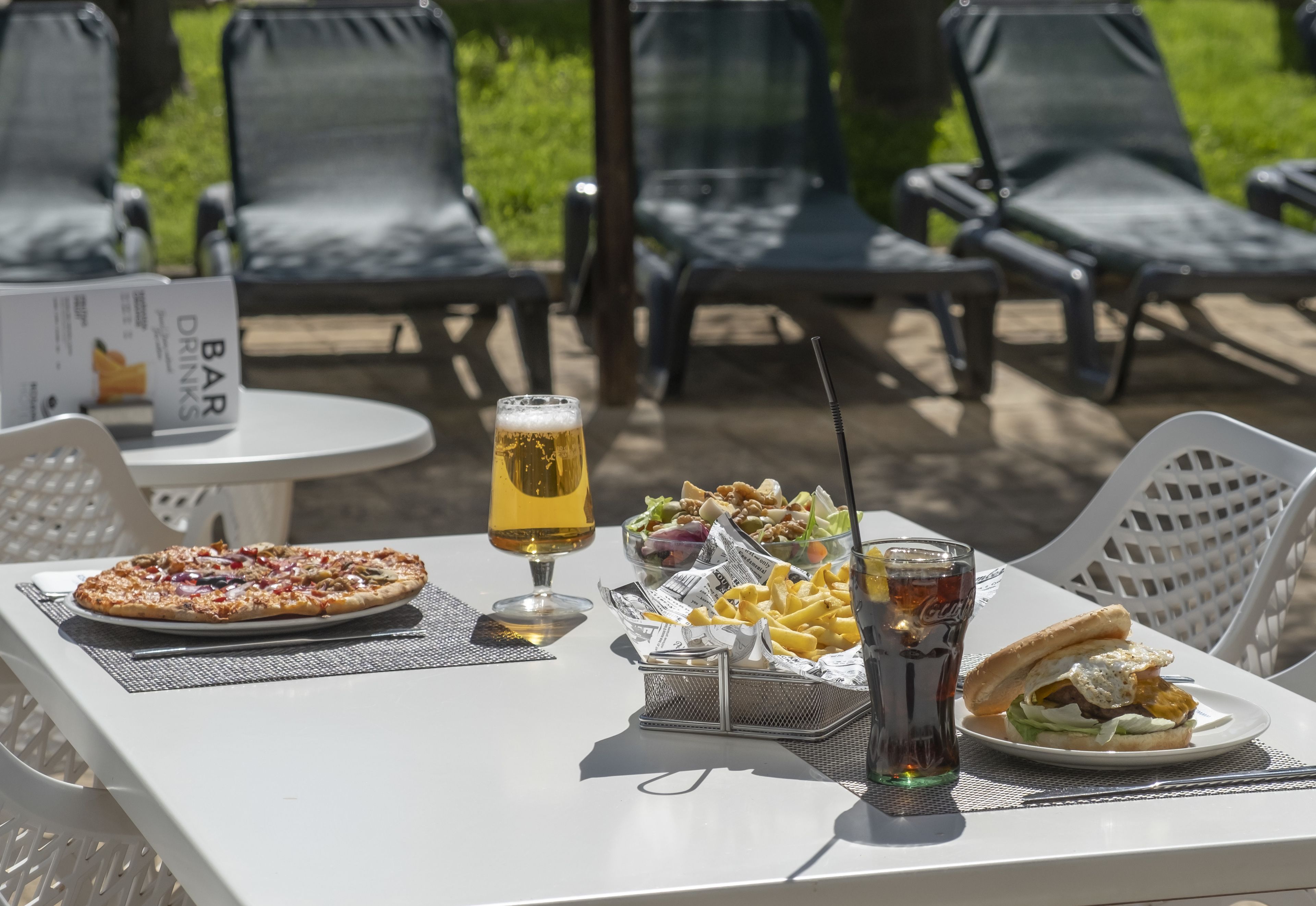 Ein Tisch mit einer Pizza, einem Salat, einem Sandwich, einem Getränk und einer Speisekarte mit der Aufschrift „BAR DRINKS“ in einer sonnigen Umgebung im Freien, im Hintergrund sind Stühle zu sehen.