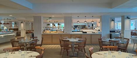 A spacious cafeteria with multiple dining tables and chairs, a central serving area, and a view of the kitchen in the background.