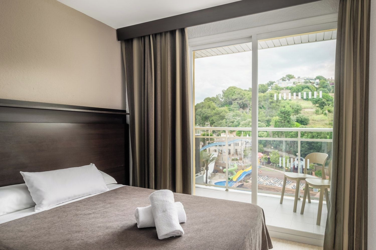 Une chambre d’hôtel avec un lit, une vue sur la piscine et l’espace extérieur, ainsi qu’un balcon avec des chaises.