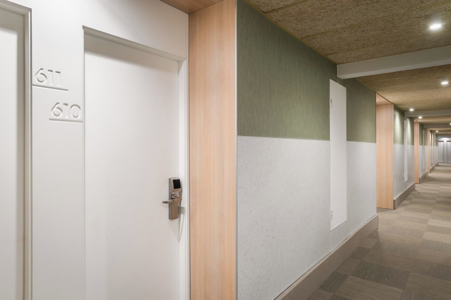 Un couloir d’hôtel avec des portes portant les numéros des chambres, un luminaire moderne et un motif de sol à carreaux.