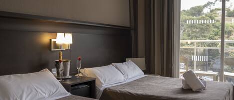 A hotel room with two beds, a nightstand, a lamp, and a view of the outdoors.