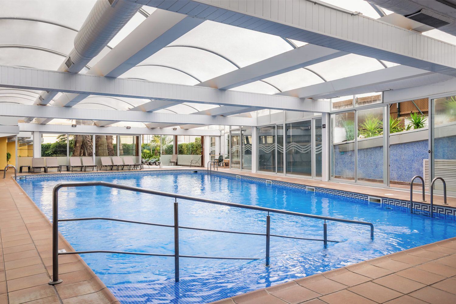 Una piscina interior con paredes de vidrio transparente, suelo de baldosas y escalera metálica.