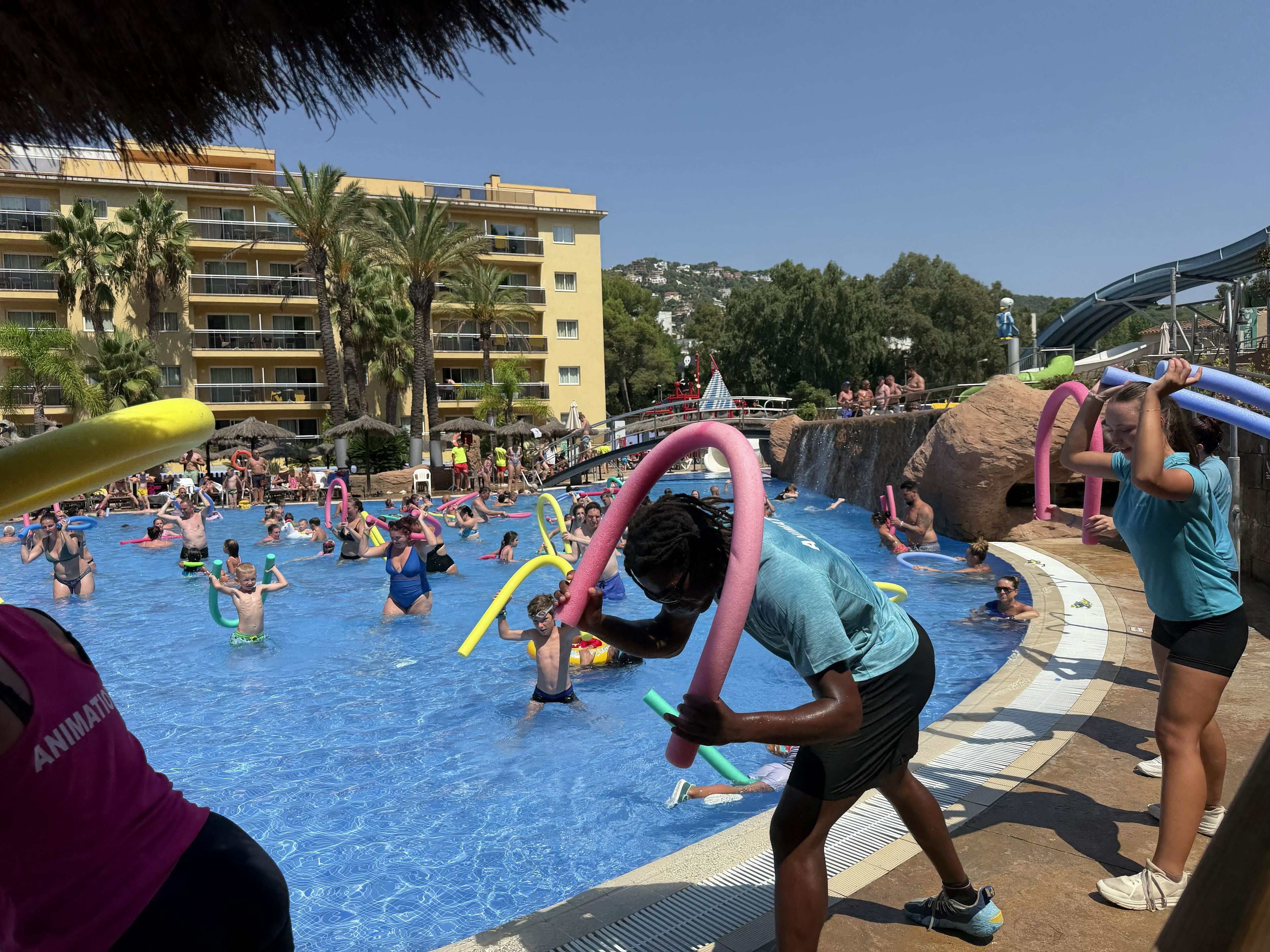 A swimming pool area with multiple levels of buildings in the background, a water slide, and people engaging in water activities.