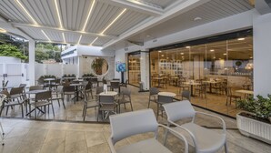 Un área de café al aire libre con mesas y sillas, una pantalla de televisión y un muro de vidrio que da a un comedor interior.