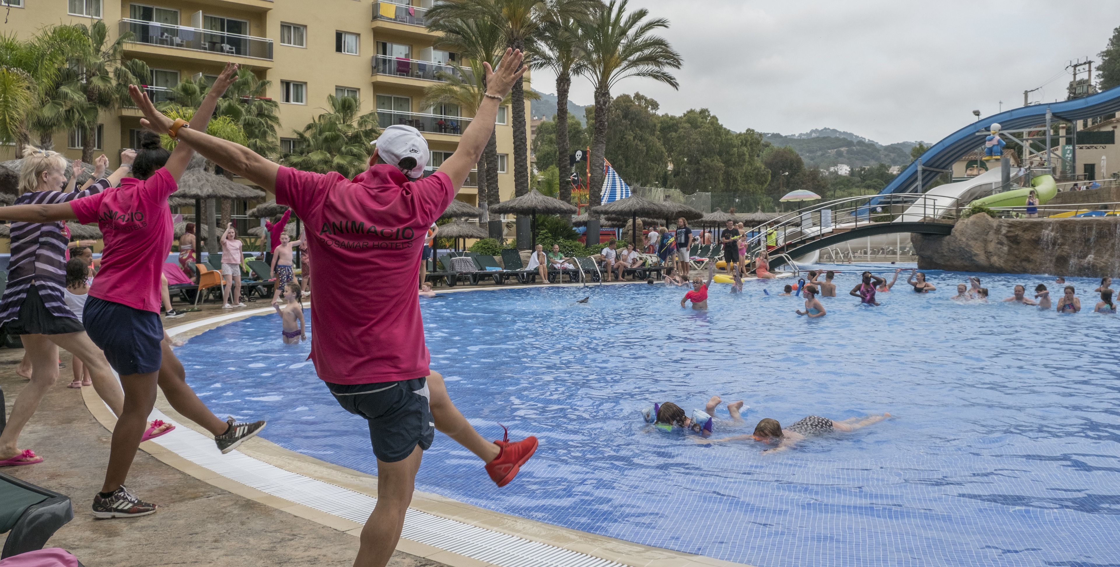 Ett poolområde där folk badar och leker, en vattenrutschkana och en hotellbyggnad i bakgrunden.