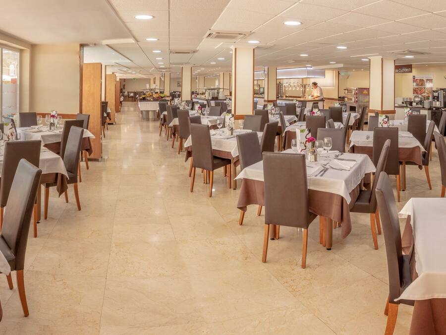 A spacious dining area with multiple tables and chairs, a kitchen in the background, and large windows offering a view.