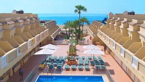 Un espace piscine d’hôtel avec de nombreux chaises longues et parasols, un palmier et une vue sur l’océan.