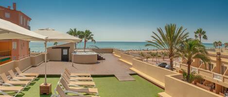 A rooftop lounge area with sun loungers, a hot tub, and a view of the beach.